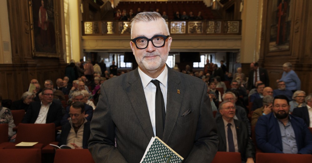 Peter De Wilde met bril en een boek in de hand in een volle zaal