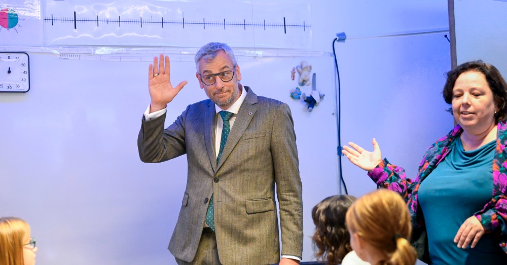 Dirk De Smedt steekt zijn hand op voor een schoolbord, naast een lachende vrouw